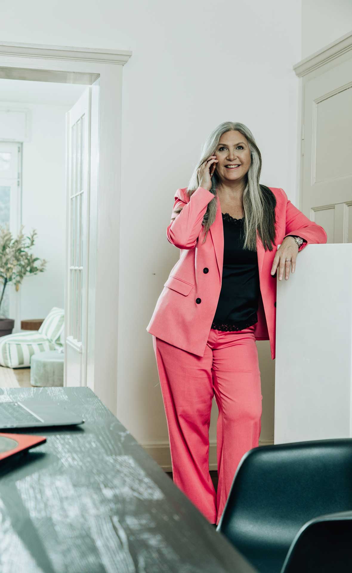 Jasminka Hepperle als Geschäftsfrau in einen rosafarbenen Anzug und schwarzer Bluse. Mit der rechten Hand hält sie ein Telefon ans Ohr und lehnt mit dem linken Arm auf ein weißes Highboard.